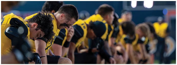 A group of people wearing yellow and black sports uniforms are kneeling in a line on a field, appearing focused and preparing for an event. Descripci n generada con IA