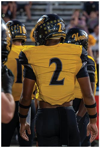 A person wearing a yellow and black football uniform with the number two on the back and a black helmet decorated with skull and crossbones stickers and the words “Sunrise Drive" is on a sports field. Descripci n generada con IA