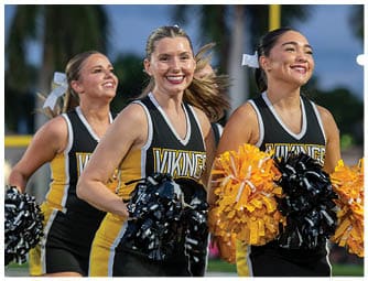 Three adults in matching black and yellow cheerleading uniforms with “VIKINGS" on the front are smiling and holding black and yellow pom-poms while performing outdoors. Descripci n generada con IA
