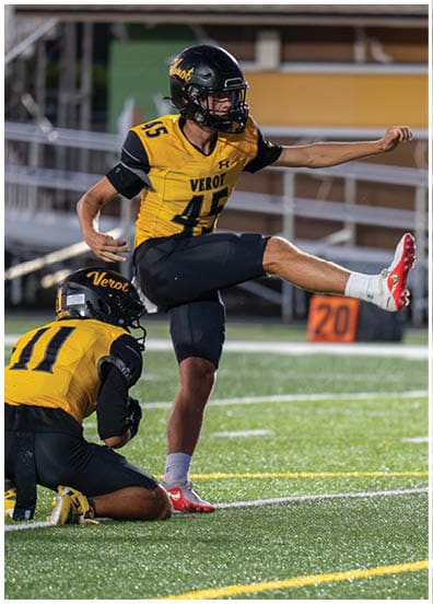 Two people in yellow and black football uniforms are on a field with one holding the ball and the other kneeling to assist with a kick while a third person in white stands nearby. Descripci n generada con IA