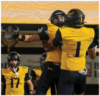Two people in yellow and black football uniforms are celebrating on a field while two others stand in the background. Descripci n generada con IA