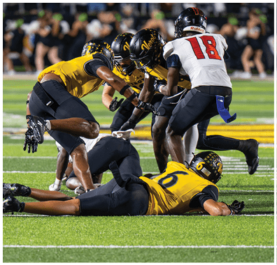 Several people wearing sports uniforms and cleats are engaged in a close physical play on a football field. Descripci n generada con IA