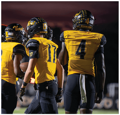Two people wearing yellow and black football uniforms with helmets stand side by side on a field at dusk. Descripci n generada con IA