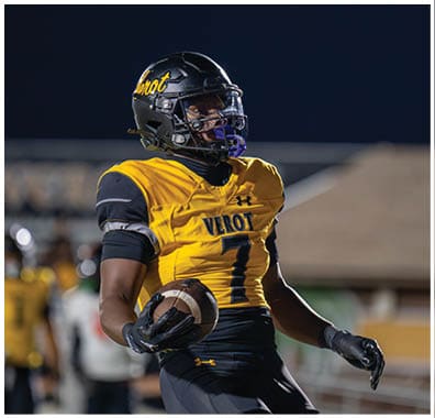 A person wearing a yellow and black football uniform with the word “VEROT" and the number seven is holding a football on a field at night. Descripci n generada con IA