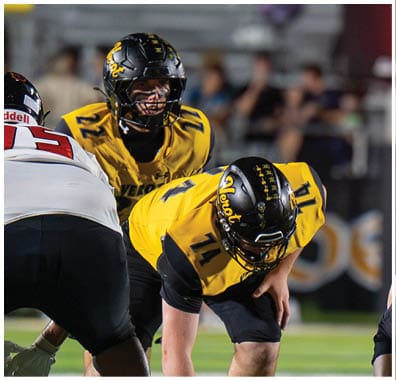 Two people in yellow and black football uniforms and helmets are positioned on a field, preparing for a play during a game. Descripci n generada con IA