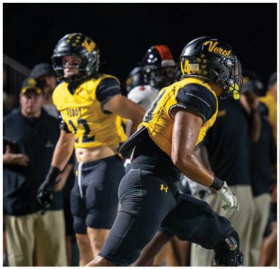Two people wearing yellow and black football uniforms and helmets are on a field at night, with one person in the foreground appearing ready to move. Descripci n generada con IA