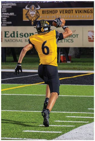 A person wearing a yellow and black football uniform with the number six is running on a green football field while holding a football in one hand. Descripci n generada con IA