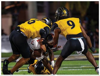 Two football players in yellow and black uniforms tackle a player in a white uniform holding a football during a game. Descripci n generada con IA