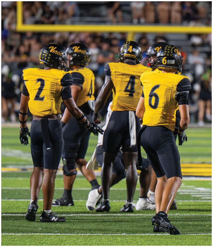 Four football players in yellow and black uniforms stand on a field with a blurred crowd in the background. Descripci n generada con IA