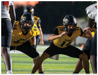 Two people in yellow and black football uniforms and helmets are crouched in a ready stance on a football field. Descripci n generada con IA