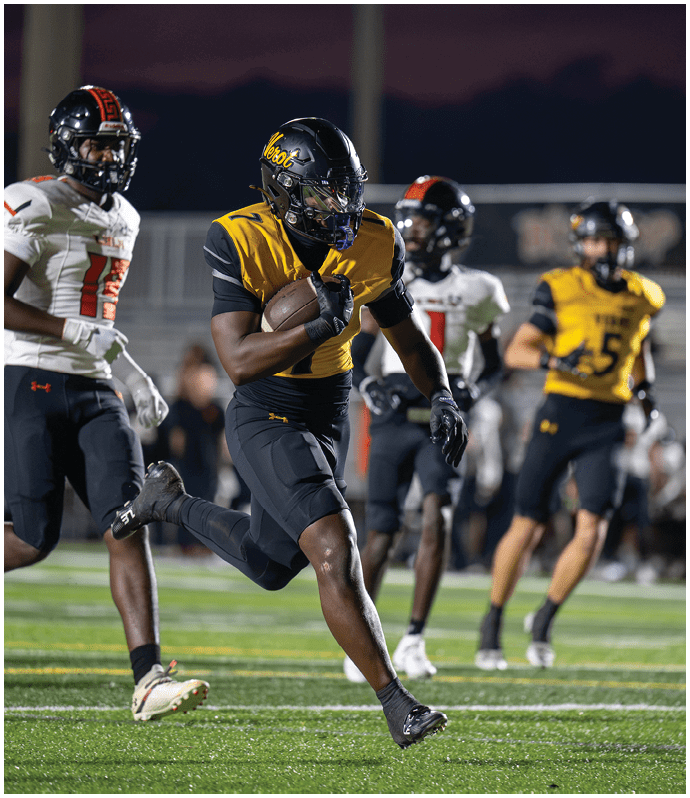 A person in a yellow and black football uniform is running with the ball on a field while other players in white and black uniforms chase behind. Descripci n generada con IA