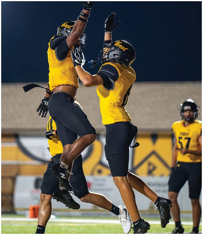 Two people in yellow and black football uniforms and helmets are celebrating by jumping and raising their arms on a field at night. Descripci n generada con IA