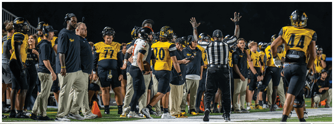 People in football uniforms and coaches stand on the sidelines of a football field at night while a referee raises both hands signaling a play. Descripci n generada con IA