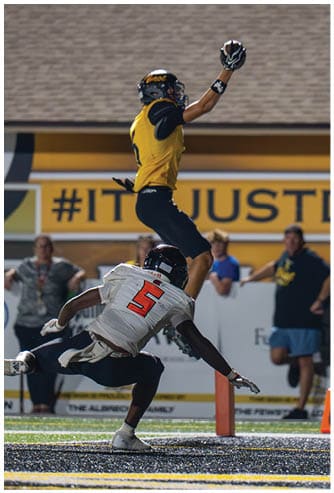 A person in a yellow and black football uniform is jumping with one arm raised holding a football while another person in a white uniform is crouched below. Descripci n generada con IA