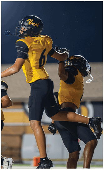 Two people in yellow and black football uniforms with helmets are celebrating on a field, with one person jumping and the other reaching up to meet them. Descripci n generada con IA