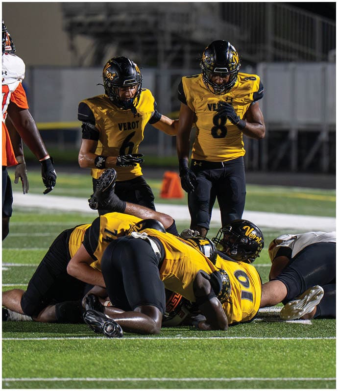 Several football players in yellow and black uniforms are engaged in a play on a field under stadium lights. Descripci n generada con IA