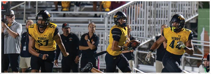 Three people in yellow and black football uniforms are actively playing on a field with spectators and staff in the background. Descripci n generada con IA