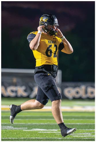 An adult wearing a yellow and black football uniform with the number 61 is adjusting their helmet while walking on a football field at night. Descripci n generada con IA