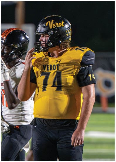 Two people in football uniforms stand on a field, one wearing a yellow and black jersey with the number 74 and the other in a white and black jersey with orange accents. Descripci n generada con IA