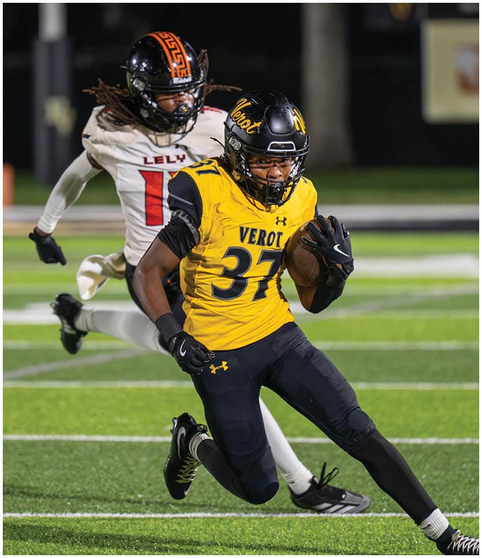 A person wearing a yellow and black football uniform is running with the ball while another person in a white and black uniform chases behind on a football field. Descripci n generada con IA