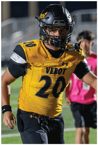 A person wearing a yellow and black football uniform with the number 20 is on a football field with another person in a pink shirt and black shorts in the background. Descripci n generada con IA