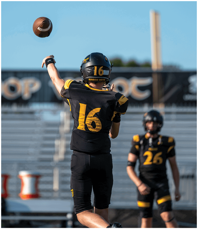 A person wearing a black and yellow football uniform with the number 16 on the helmet is throwing a football against a clear blue sky. Descripci n generada con IA