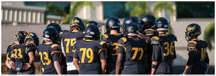 A group of people wearing black and yellow football uniforms and helmets are gathered closely together outdoors, likely during a team huddle. Descripci n generada con IA