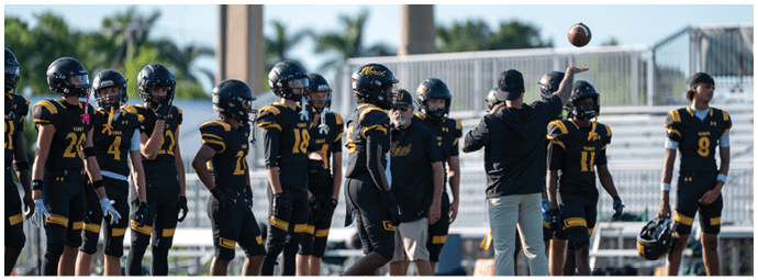 A group of people wearing black and yellow football uniforms stand on a field while one person in casual clothing tosses a football in the air. Descripci n generada con IA