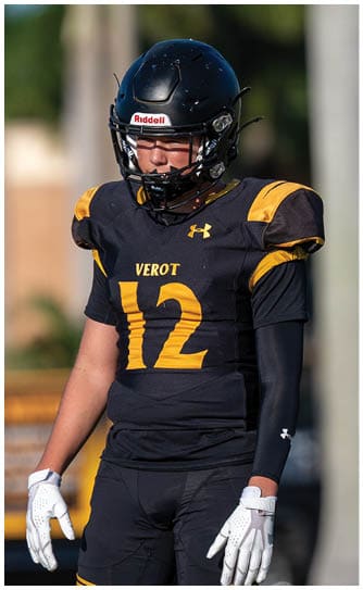 A person wearing a black and yellow football uniform with the number 12 and a helmet stands outdoors with a blurred background of greenery and structures. Descripci n generada con IA