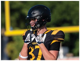 A person wearing a black and yellow football uniform and helmet is adjusting their gloves on a field with a blurred goalpost and trees in the background. Descripci n generada con IA