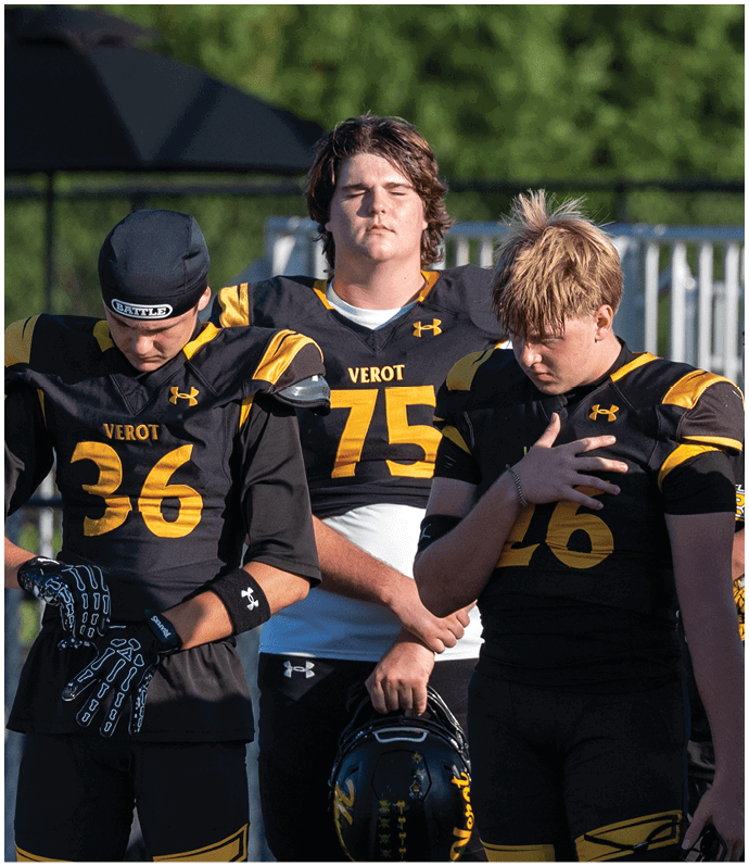 Three people wearing black and yellow football uniforms stand with their heads bowed, one holding a helmet and another with a hand on their chest. Descripci n generada con IA