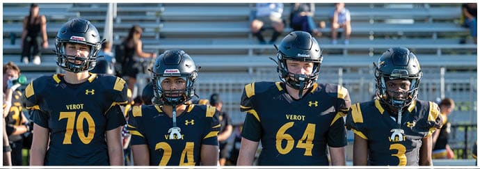 Four people wearing black and yellow football uniforms with helmets stand side by side on a sports field with bleachers in the background. Descripci n generada con IA