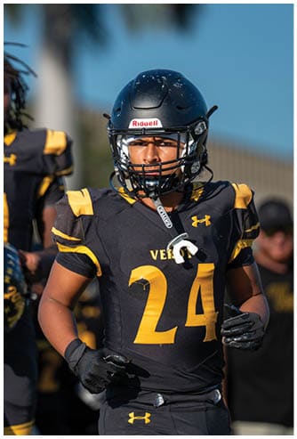 A person wearing a black and yellow football uniform with the number 24 and a black helmet is standing outdoors with a focused expression. Descripci n generada con IA
