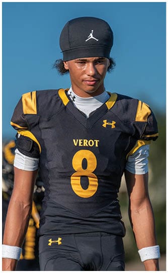 A person wearing a black and yellow sports jersey and a black cap stands against a clear blue sky. Descripci n generada con IA