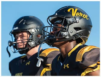 Two people wearing black football helmets and yellow and black uniforms are standing outdoors against a clear blue sky. Descripci n generada con IA