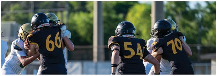 People wearing black and gold football uniforms are engaged in a play against opponents in white and gold uniforms on a grassy field with trees in the background. Descripci n generada con IA