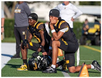 Two people in black and yellow football uniforms kneel on a field with helmets on the ground near orange cones while others stand in the background. Descripci n generada con IA