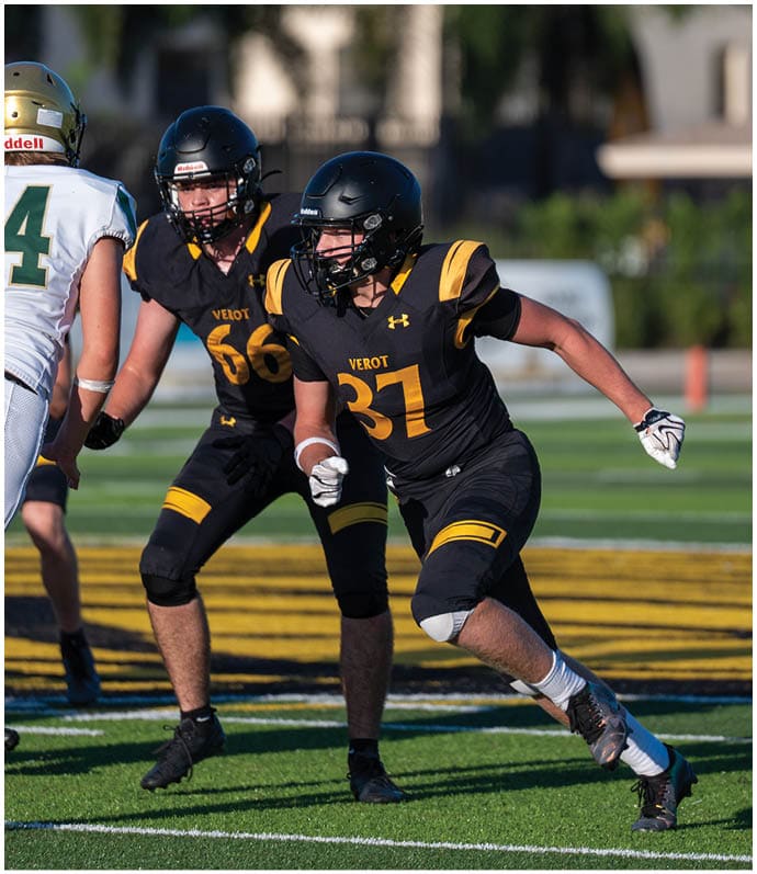 Two people in black and yellow football uniforms are running on a field toward a person in a white and green uniform during a game. Descripci n generada con IA
