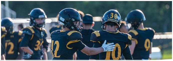 A group of people wearing football helmets and uniforms are gathered outdoors, with one person placing a hand on another's shoulder. Descripci n generada con IA