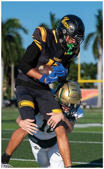 A person wearing a black and yellow football uniform with a helmet is holding a football tightly while another player in a gold helmet is partially visible below. Descripci n generada con IA