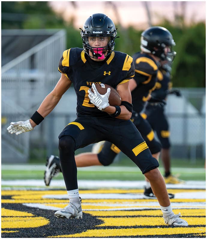 A person in a black and yellow football uniform is running on a field while holding a football, with another player in similar attire in the background. Descripci n generada con IA