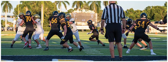 Adults in football uniforms are engaged in a game on a grassy field with a referee standing near the center. Descripci n generada con IA
