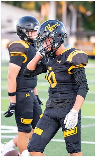 Two people in black and yellow football uniforms and helmets stand on a field with one person adjusting their helmet. Descripci n generada con IA