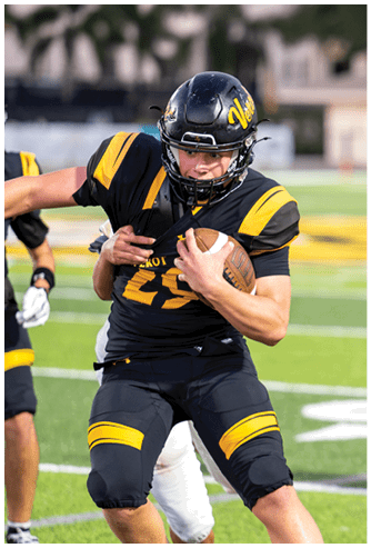 An adult wearing a black and yellow football uniform is running with a football while being tackled by another player on a green field. Descripci n generada con IA