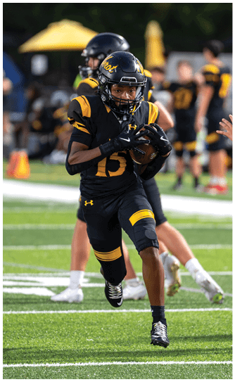 A person in a black and yellow football uniform is running on a field while holding a football, with other players and a hand reaching toward them in the background. Descripci n generada con IA