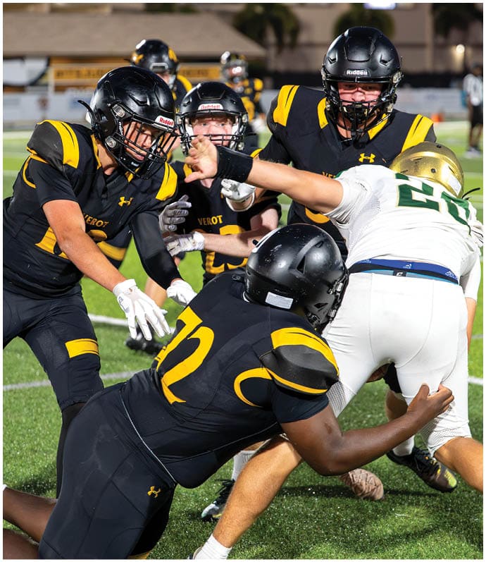 Four people in football uniforms are actively engaged in a play on a green field with one person in white attempting to move past three people in black and yellow uniforms. Descripci n generada con IA