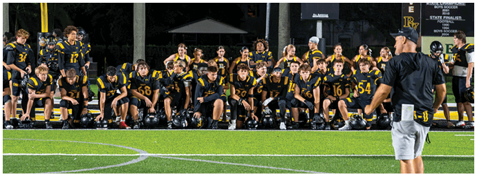 A group of people in black and yellow sports uniforms sit and stand on the sidelines of a football field while a person in a black shirt and gray shorts stands facing them. Descripci n generada con IA