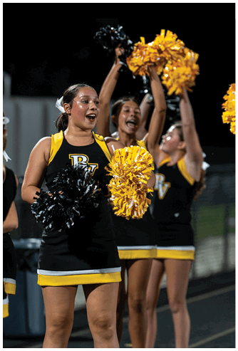 A person in a black and yellow cheerleading uniform is holding black and yellow pom-poms and appears to be cheering with others in similar uniforms in the background. Descripci n generada con IA