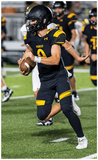 A person in a black and yellow football uniform is running on a field while holding a football, with other players visible in the background. Descripci n generada con IA