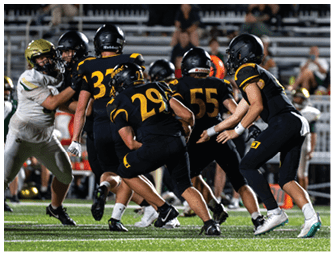 A group of people wearing black and yellow football uniforms are engaged in a play on a green field while people in white and gold uniforms attempt to block them. Descripci n generada con IA